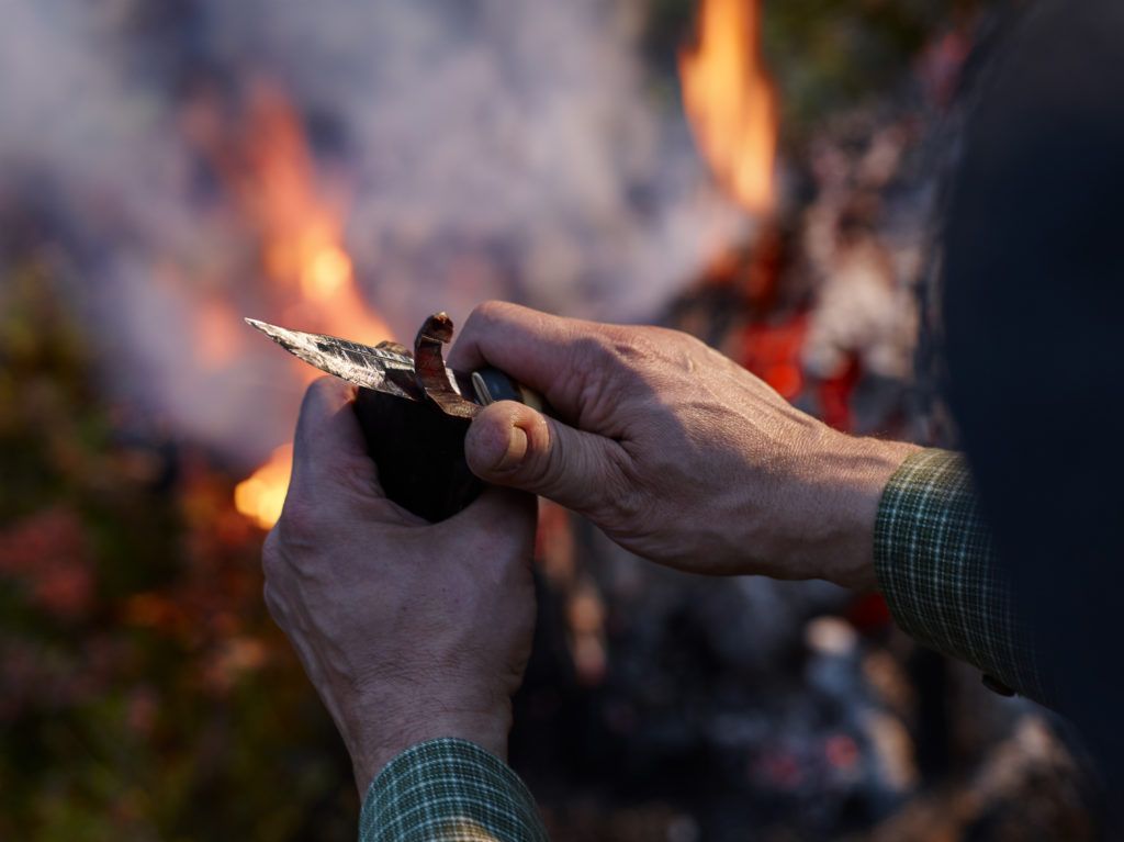 Mann som skjærer speket reinskjøtt med kniv. Bål i bakgrunn. 