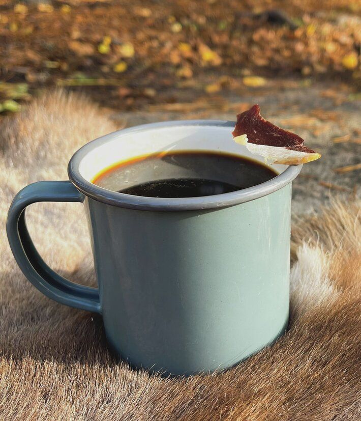 Grønn aluminiumskopp med kaffe. På kanten av koppen hviler en bit med tørket reindykjøtt. 